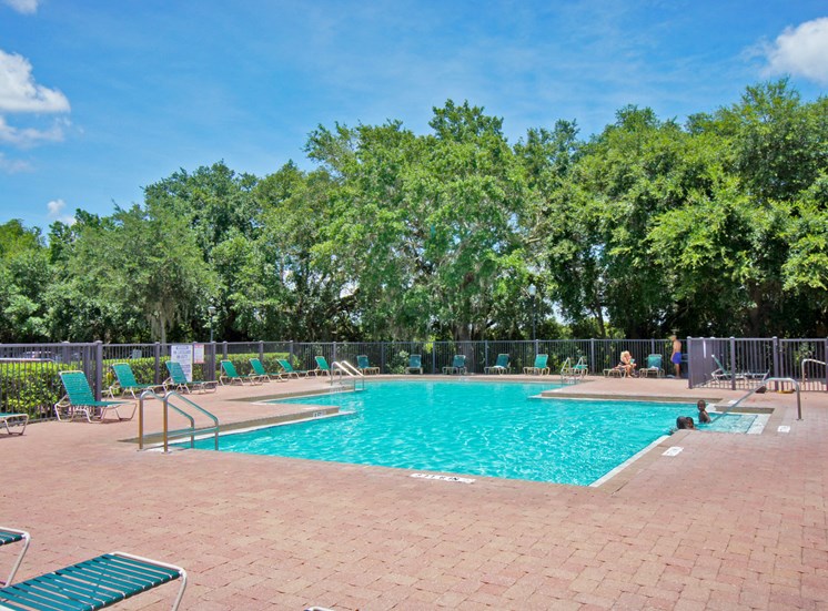 Community pool with sundeck, lounge chairs, surrounded by grey fence with trees, in the background